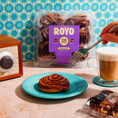 A hand holding a fork reaches for a cinnamon roll on a turquoise plate, with a package of Royo cinnamon rolls and a cup of coffee in a cozy kitchen setting featuring a patterned background.
